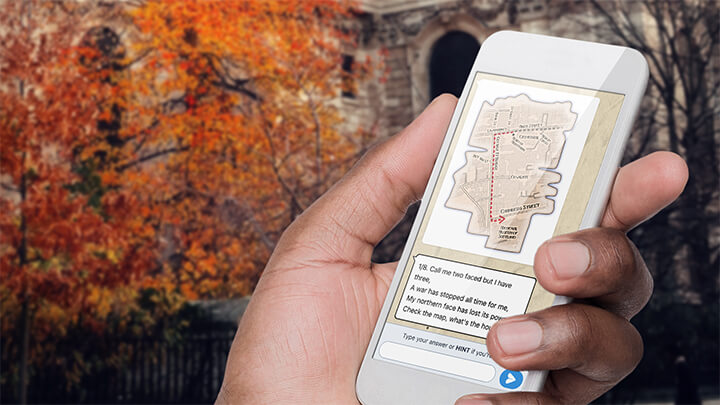 A hand holding a phone playing Treasure Hunt Birmingham with a tree with orange leaves behind.