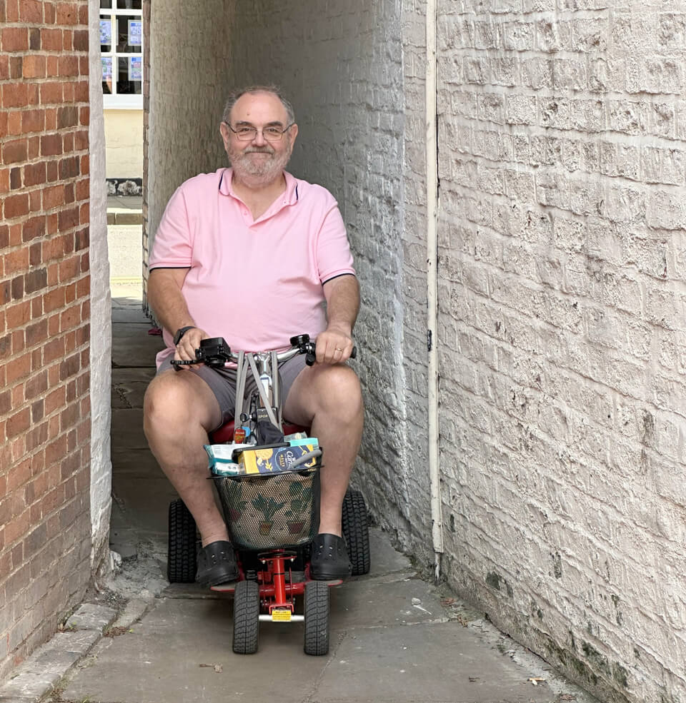 A man riding a small mobility scooter on a treasure hunt, emerging from a narrow alleyway with white brick walls on one side and red brick on the other. The alleyway is just wide enough to fit the scooter